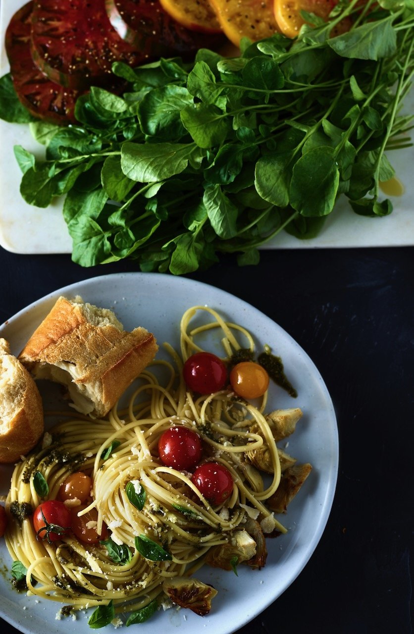 A plate of spaghetti with cherry tomatoes, basil, grated cheese, and bread on the side, with a bunch of fresh green arugula and heirloom tomatoes on the cutting board in the background.
