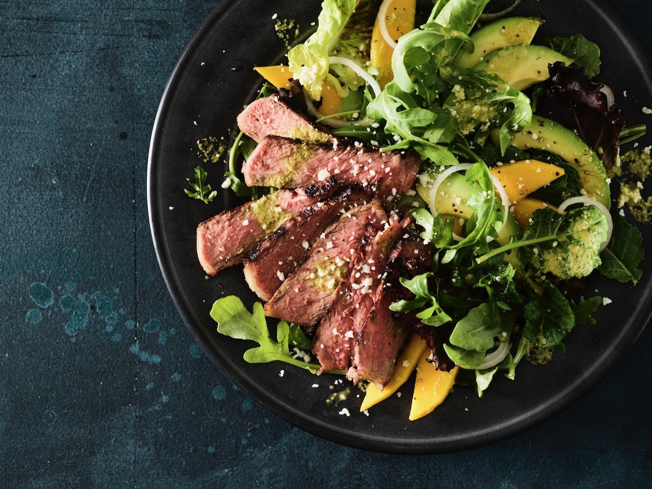 A black plate containing seared steak slices on a bed of mixed greens, sliced avocado, yellow bell peppers, and sprinkled cheese, on a dark textured surface. Food Styled in Denver, Colorado by Beth Hawkins of Hawkins and Company