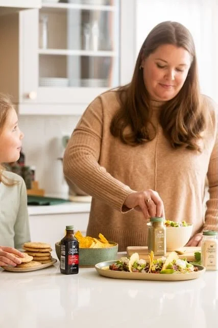 A woman and a young girl preparing food together in a kitchen, with vegetables, condiments, and a plate of salad on the counter.