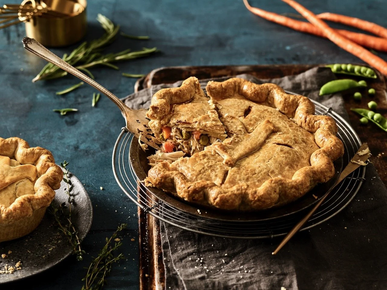 A homemade chicken pot pie with a golden crust on a wire cooling rack, with a slice removed showing vegetables and chicken filling, surrounded by carrots, green peas, and fresh herbs on a rustic dark surface. Food styled for Hinman Pies, in Denver, C