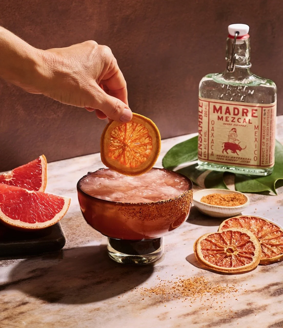 A hand places a dried orange slice into a cocktail glass filled with ice, on a marble surface with grapefruit slices, dried citrus slices, a bottle of Madre Mezcal, a bowl of orange spice, and green leaves surrounding it.