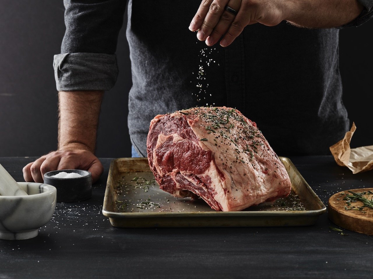 A person seasoning a large cut of raw beef with salt and herbs on a baking sheet. Food styled for Beef It's What's for Dinner in Denver, Colorado by food stylist Beth Hawkins of Hawkins and Company