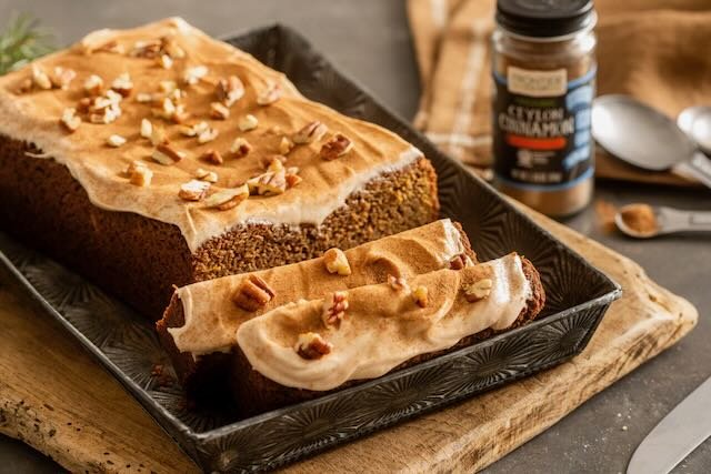 Banana bread topped with chopped nuts, placed on a black serving tray on a wooden cutting board, with a jar of cinnamon and a spoon in the background.