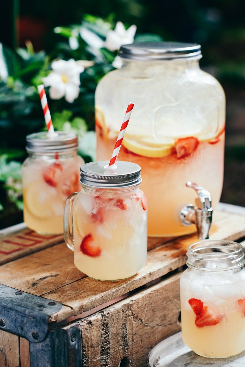 Glass jar beverages with lemon slices and strawberries, served with striped paper straws on a rustic wooden table near a large drink dispenser. In the background, white flowers and greenery are visible.