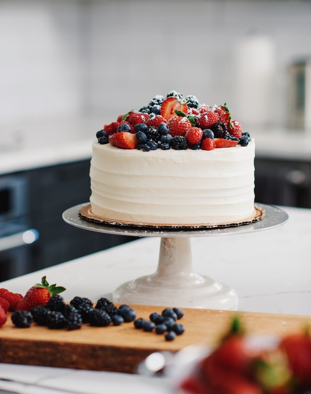 A white frosted cake topped with mixed berries on a cake stand in a kitchen.