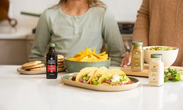 People in a kitchen with a platter of sandwiches, a bowl of potato chips, and bottles of condiments on the countertop.