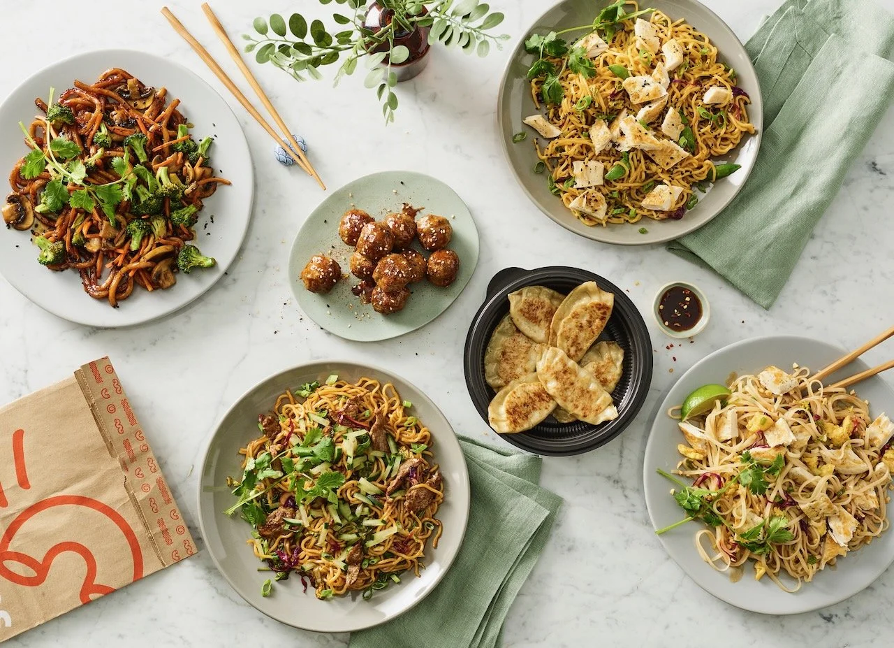 Assorted Asian dishes on a white marble table, including noodles with vegetables and chicken, dumplings, and meatballs, garnished with cilantro and green onions. Food styled for Noodles and Company in Denver, Colorado by food stylist Beth Hawkins of 