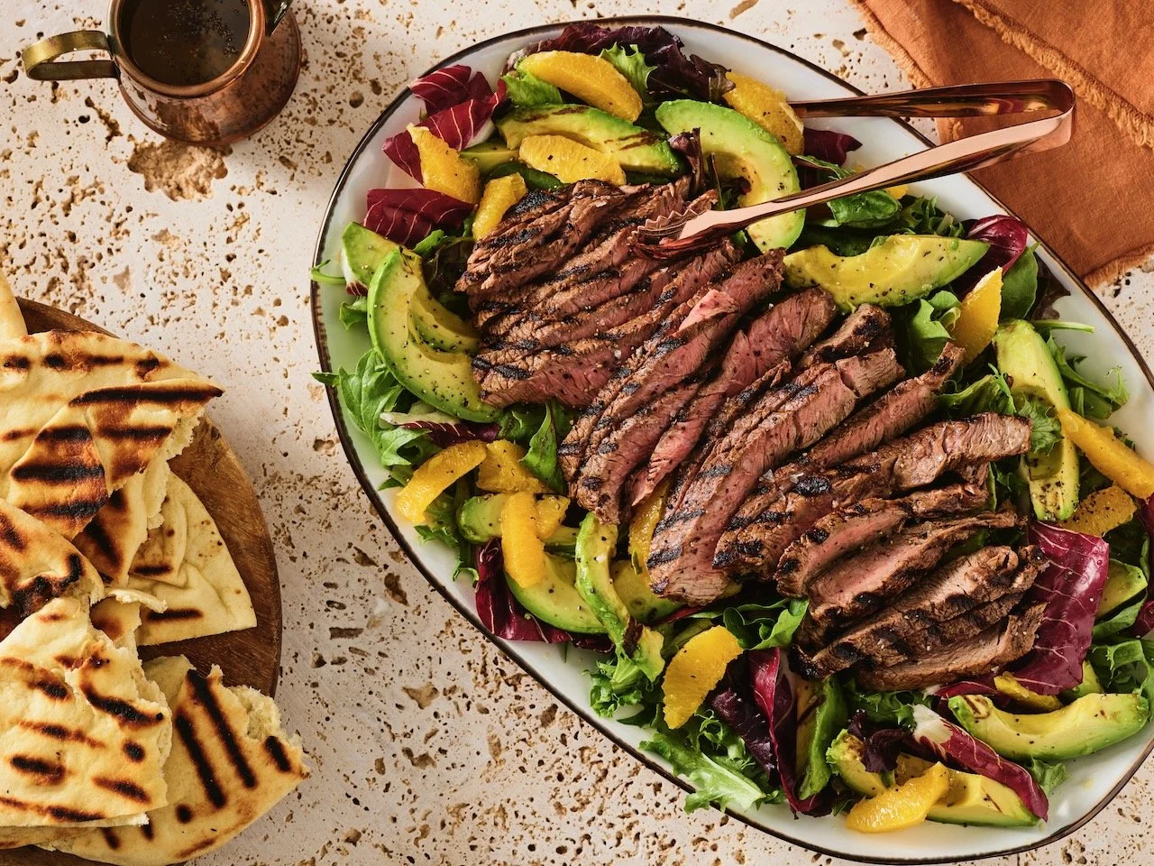 Grilled steak salad with mixed greens, avocado, yellow bell peppers, and lemon wedges, served in a white bowl with tongs. Portion of grilled flatbread on a wooden board nearby. Food Styled in Denver, Colorado by Beth Hawkins of Hawkins and Company