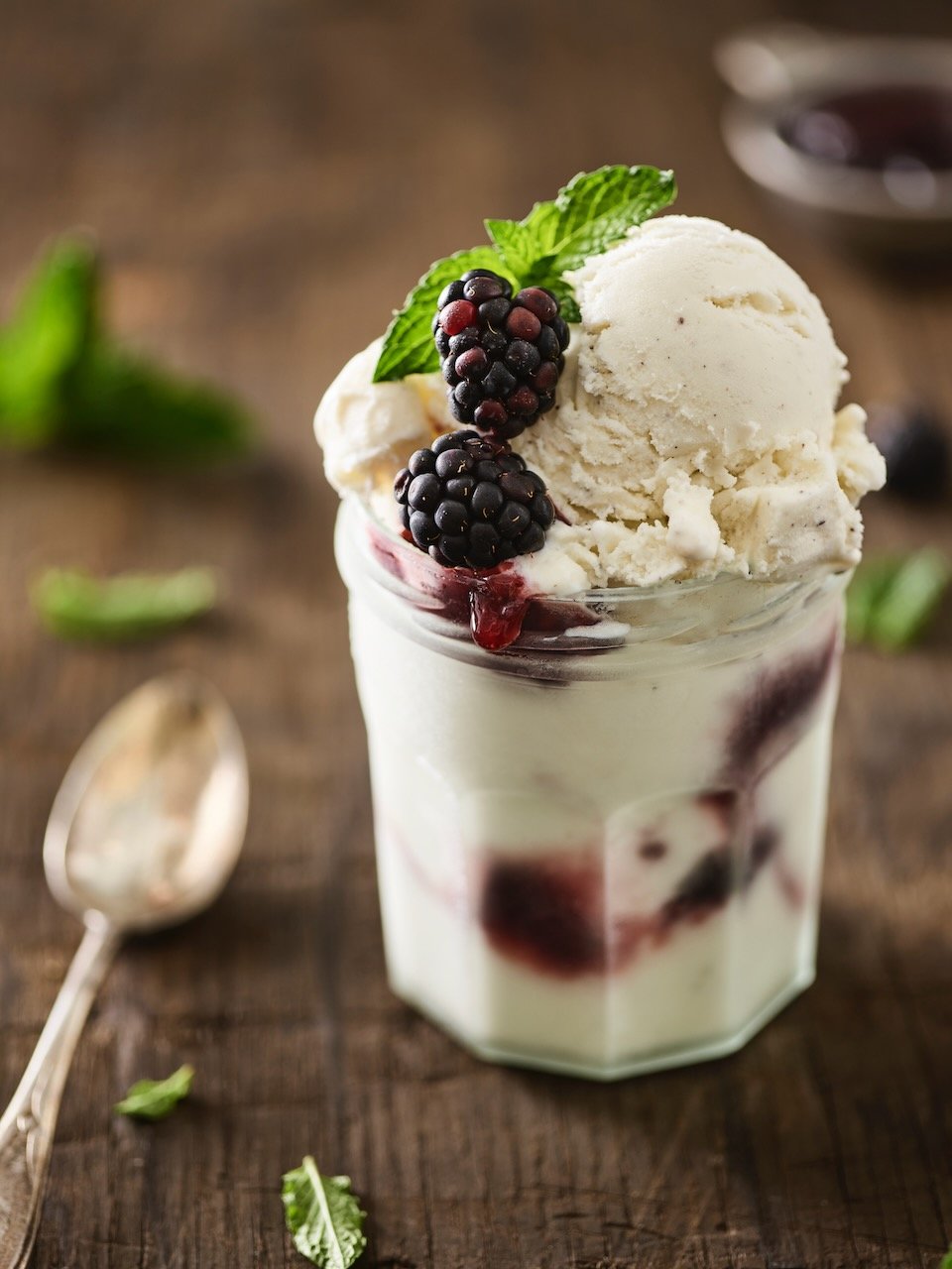 A glass of vanilla ice cream topped with blackberries and mint leaves, with berry syrup drizzled around.