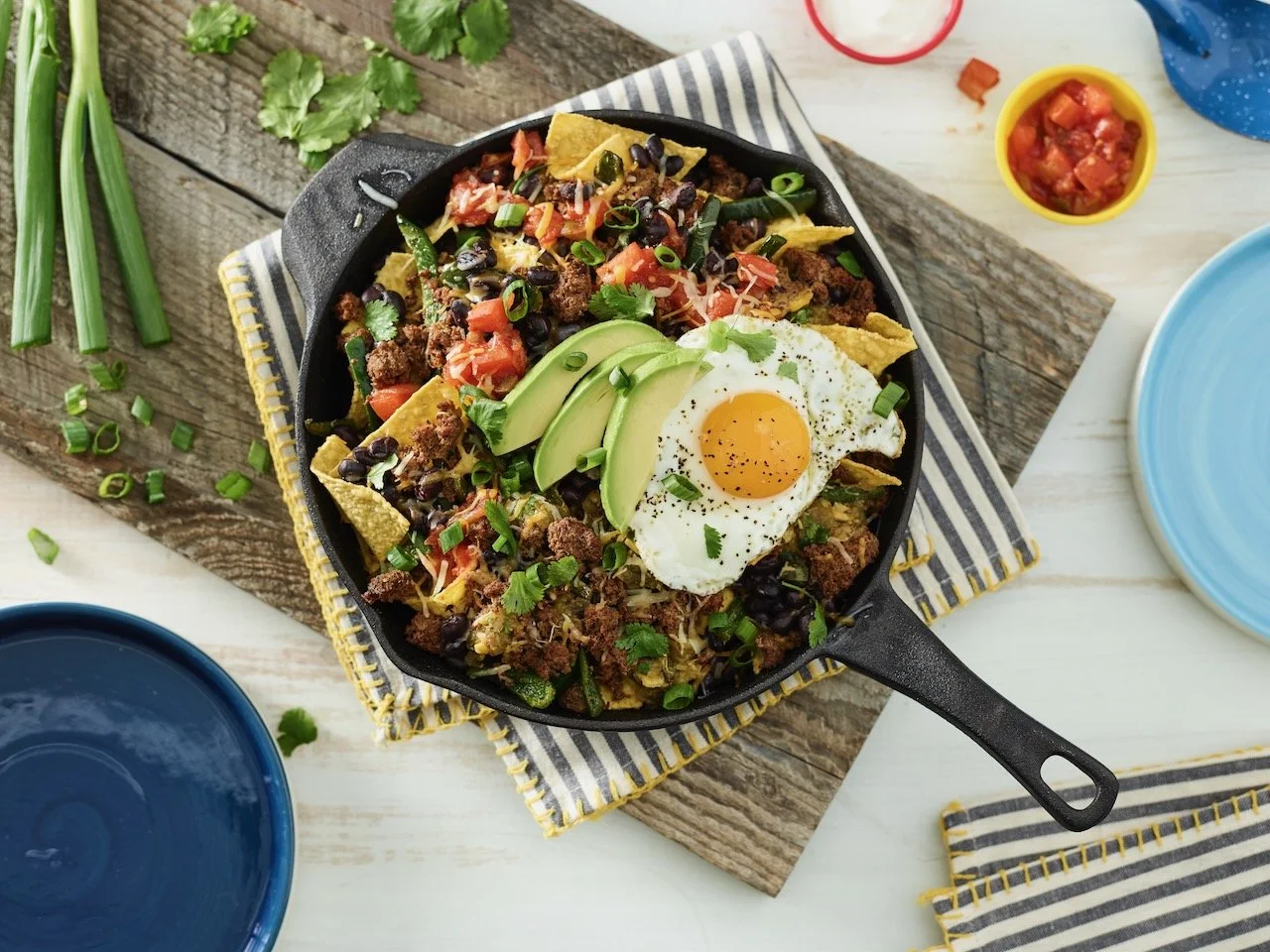 A skillet of breakfast hash topped with a fried egg, avocado slices, chives, shredded cheese, and black beans, surrounded by blue and yellow bowls, fresh cilantro, green onions, and a striped napkin on a rustic wooden surface.
