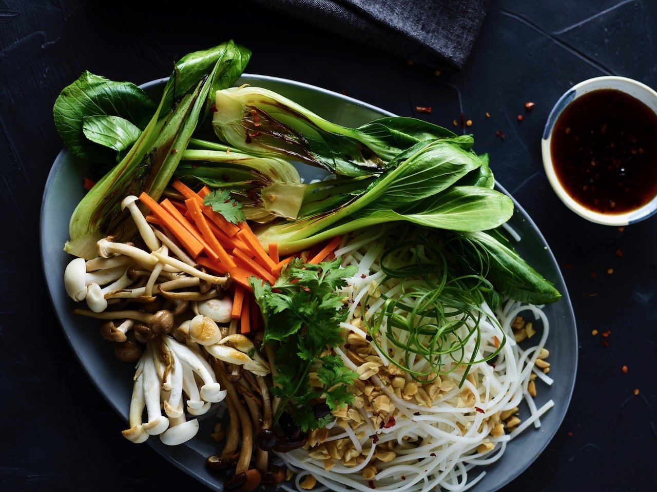 A plate of fresh vegetables including bok choy, carrots, enoki mushrooms, other various greens, and bean sprouts, with a small bowl of soy sauce on the side.