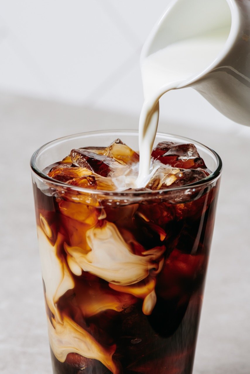 A glass of iced coffee with cream being poured from a white pitcher.