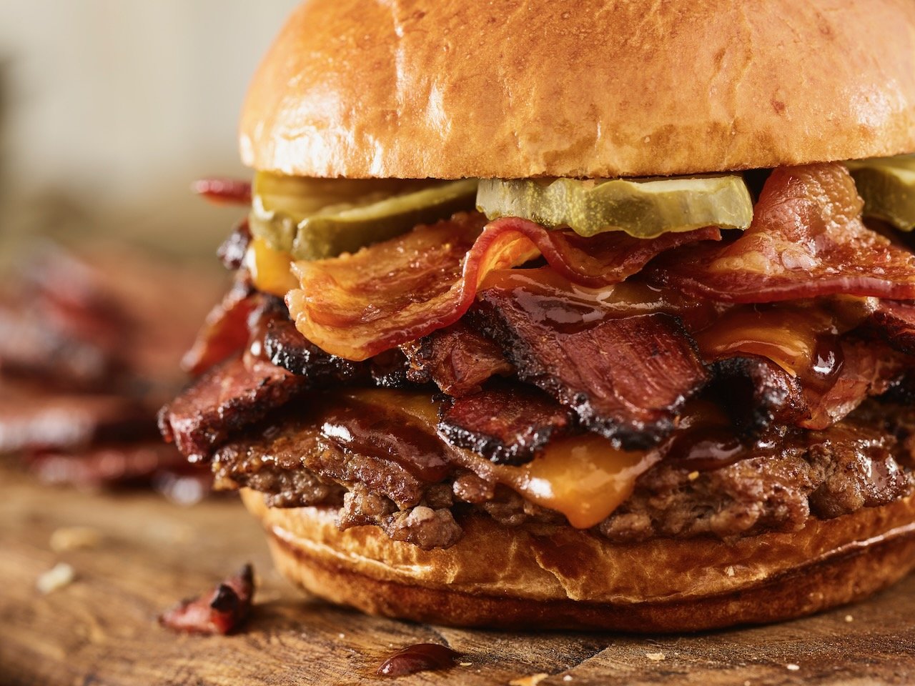 Close-up of a loaded cheeseburger with pickles, bacon, beef patty, and melted cheese on a wooden surface. Food styled for Smashburger in Denver, Colorado by food stylist Beth Hawkins of Hawkins and Company
