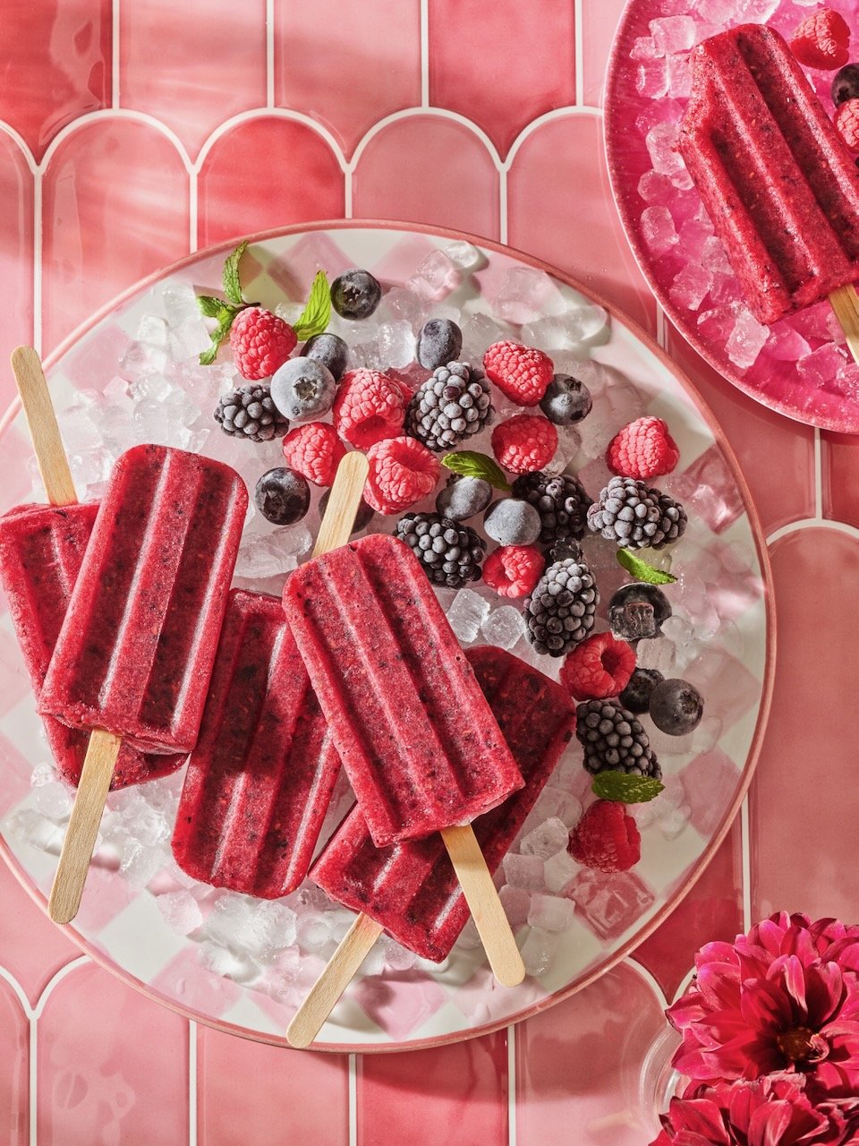 Four red berry popsicles on a plate of ice with mixed berries and green leaves, set on a pink tiled surface with additional berry popsicle and pink flowers.