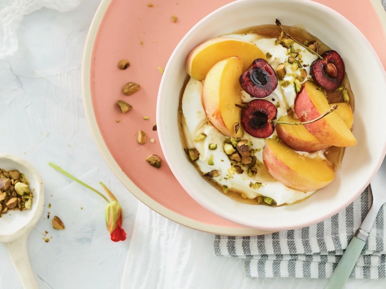 Yogurt topped with sliced peaches, cherries, and chopped pistachios in a white bowl on a pink and white tray.