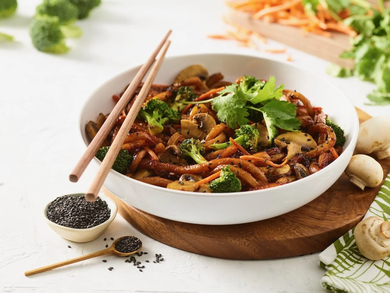 A bowl of stir-fried vegetables with broccoli, carrots, mushrooms, and cilantro, on a wooden board with black sesame seeds and chopsticks nearby. Food styled for Noodles and Company in Denver, Colorado by food stylist Beth Hawkins of Hawkins and Comp