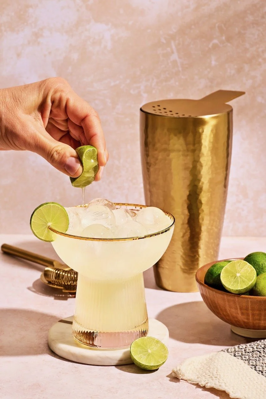 A hand squeezing lime into a glass filled with ice and a light-colored beverage, with whole and halved limes on the table. A gold cocktail shaker is in the background.