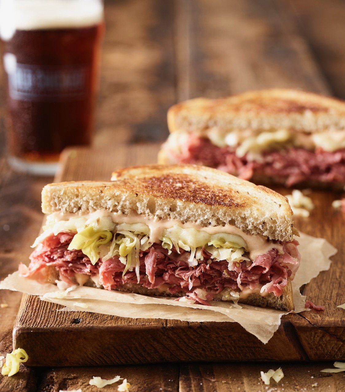 Close-up of a rueben sandwich with lettuce and sauce on toasted bread, served on a wooden board with a glass of beer in the background. Food styled in Denver, Colorado by food stylist Beth Hawkins of Hawkins and Company