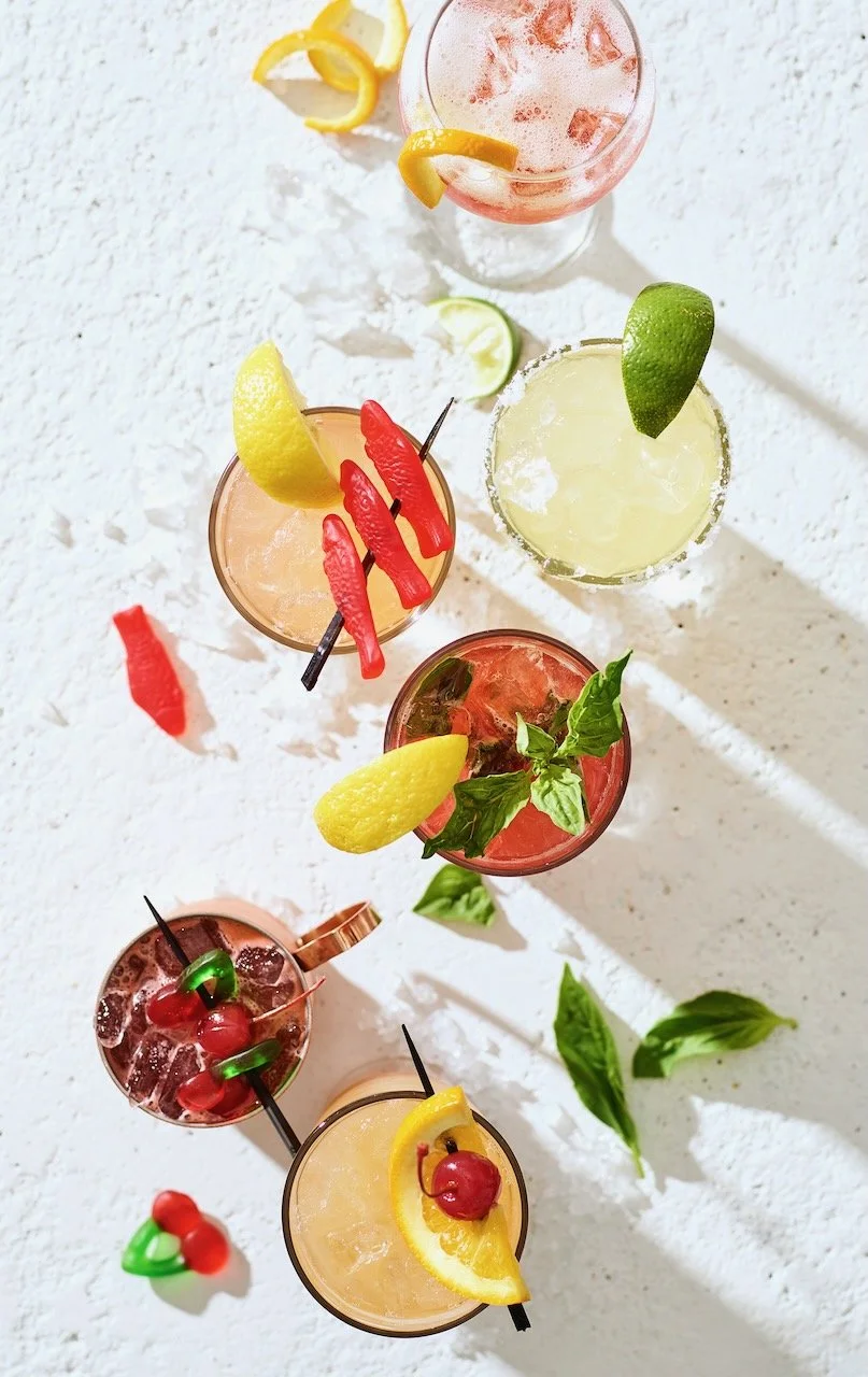 Six colorful cocktails garnished with fruit slices and herbs, arranged on a white textured surface with scattered garnishes and small fruit decorations.