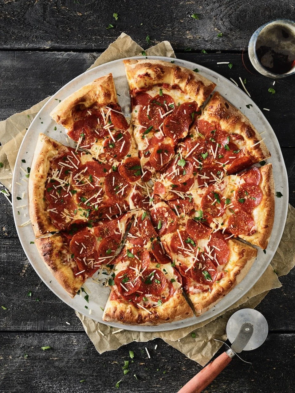A pepperoni pizza with shredded cheese and chopped herbs on a white plate, served on a dark wooden table with a glass of soda and a pizza cutter. Food styled for Old Chicago in Denver, Colorado by food stylist Beth Hawkins of Hawkins and Company