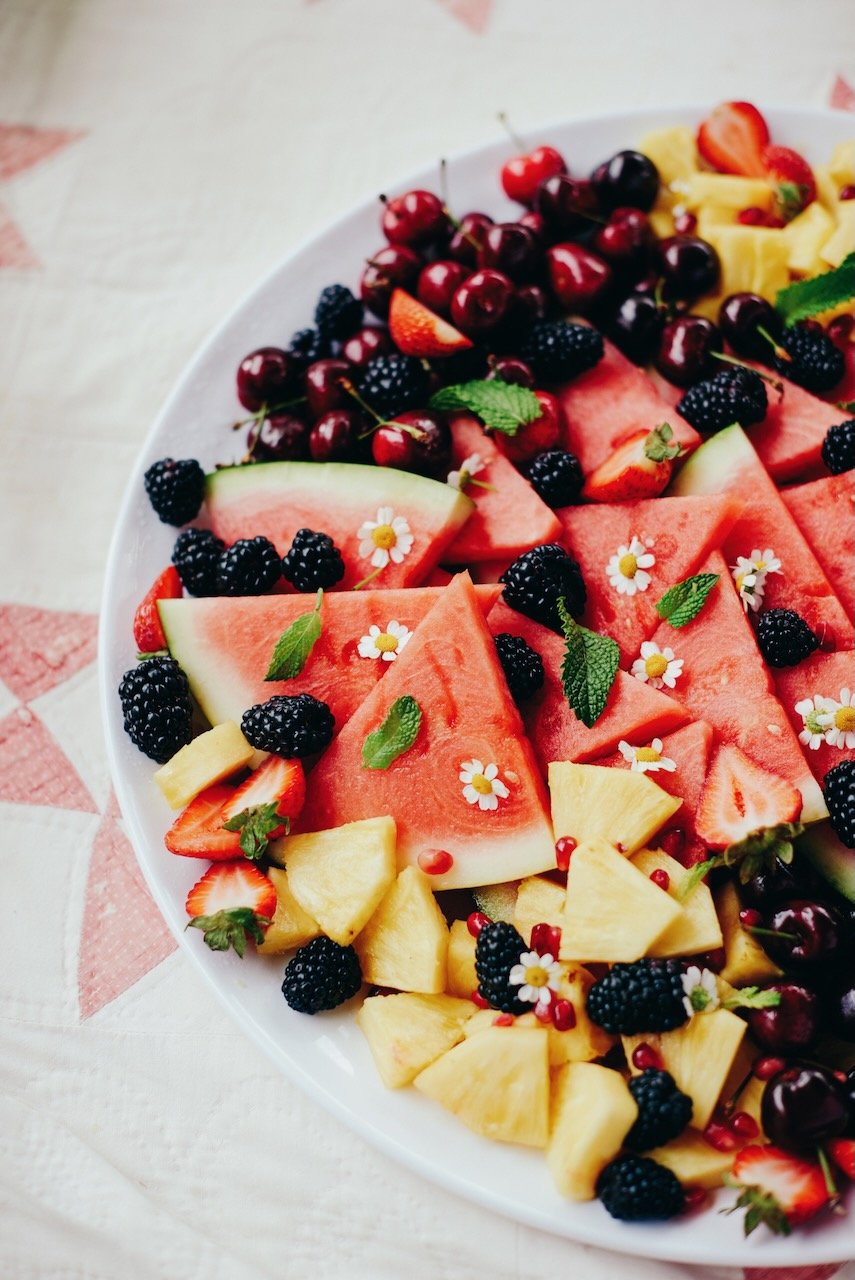 A platter of sliced watermelon, pineapple, strawberries, blackberries, cherries, and garnished with mint leaves and small white flowers.