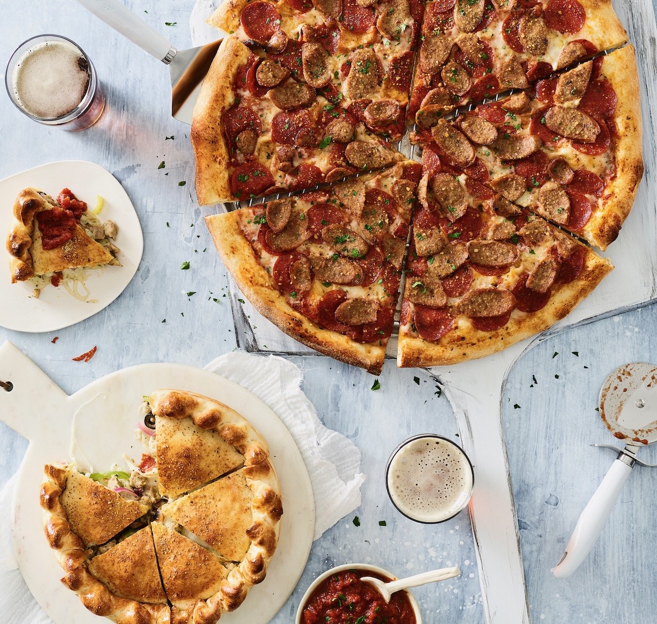 A large pepperoni and sausage pizza on a silver pizza tray with slices cut. Two smaller pizzas, one with cheese and toppings, and the other with a golden crust. Two glasses of beer, a bowl of marinara sauce, and a pizza cutter are also visible on a l