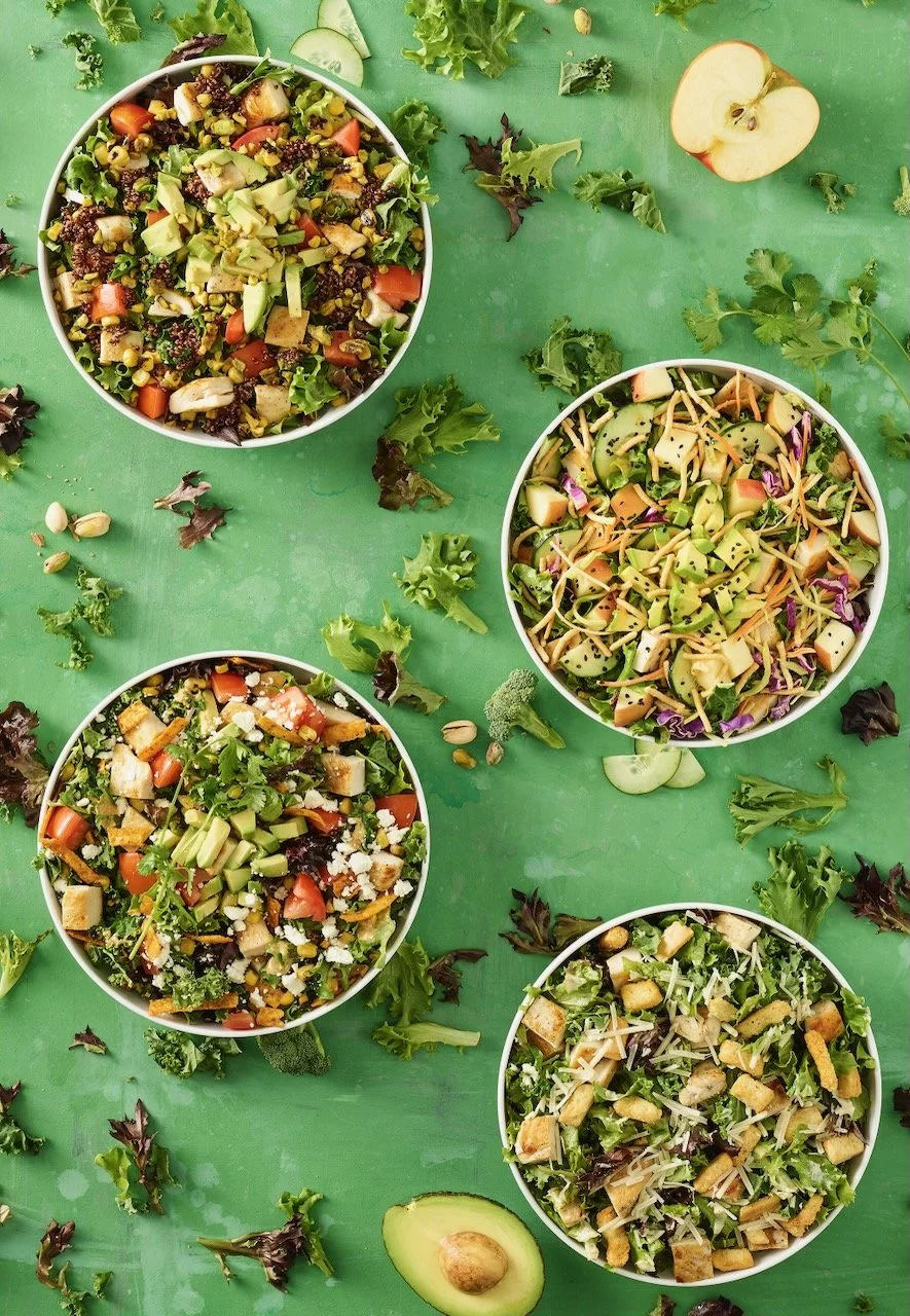 Four bowls of salad with various vegetables and toppings on a green surface, with scattered greens, cucumber slices, apple halves, and an avocado. Food Styled for Noodles and Company, in Denver, Colorado by food stylist Beth Hawkins of Hawkins and Co
