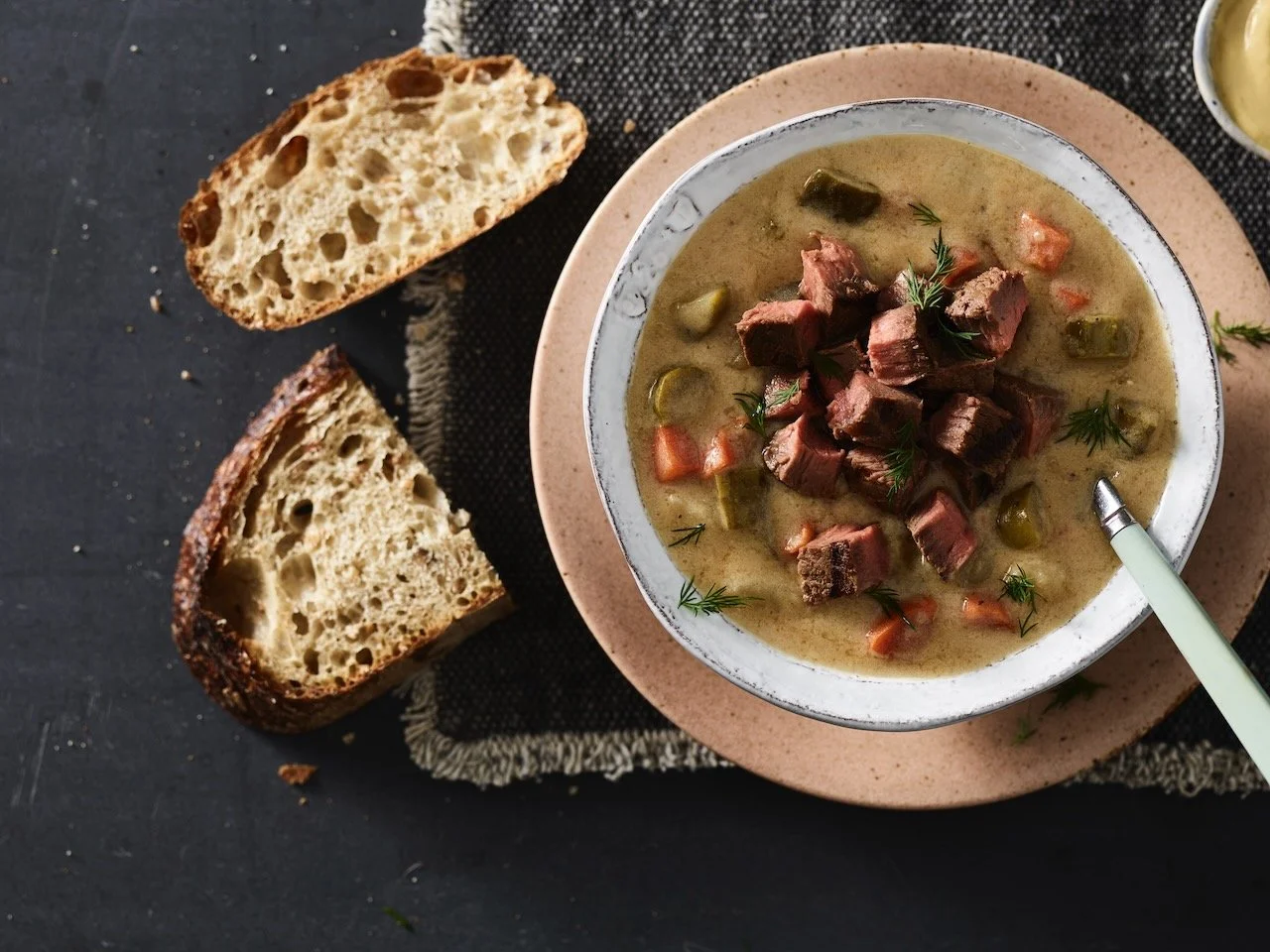 A bowl of beef stew with vegetables on a pink plate, with slices of rustic bread on a black surface. Food Stylist Colorado Food Stylist Denver. Hawkins and Company