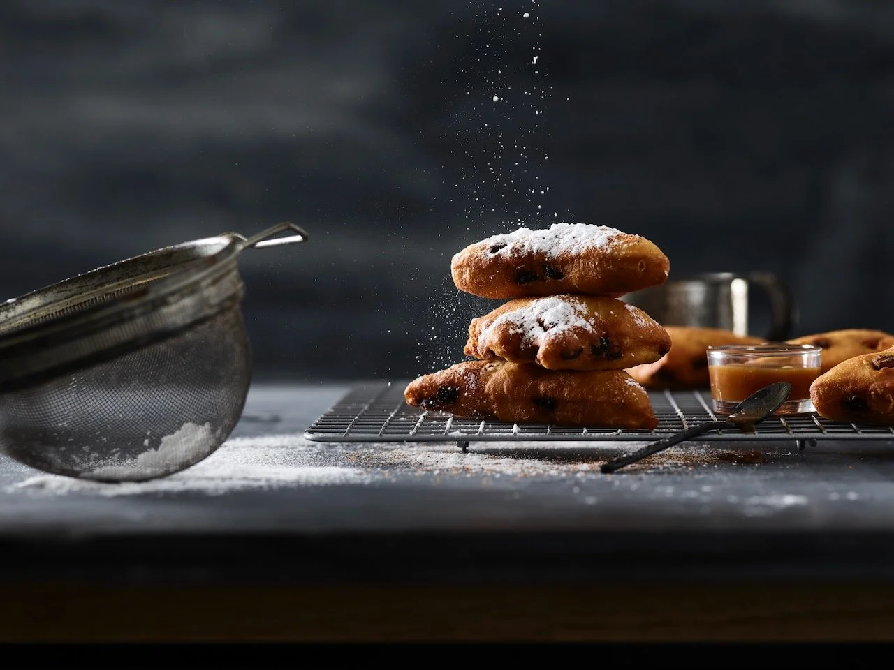 A stack of chocolate chip cookies with powdered sugar being sprinkled on top, placed on a cooling rack with a jar of caramel and a spoon beside them, and a metal sieve nearby.