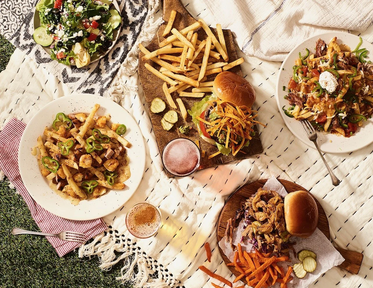 A picnic table set with various dishes including salad, French fries, pasta, burgers, and drinks on a white tablecloth. Food styled for Old Chicago in Denver, Colorado by food stylist Beth Hawkins of Hawkins and Company