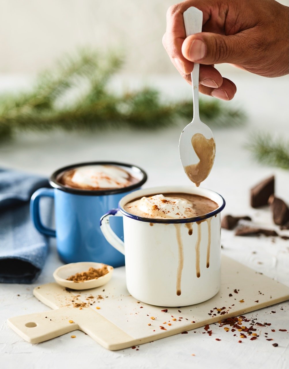 Close-up of a hand holding a spoon over a mug of hot chocolate topped with whipped cream in a white mug with a blue rim; another blue mug and scattered chocolate pieces in the background.