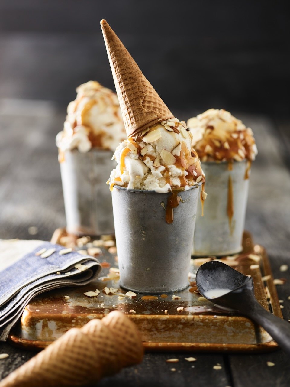 Three ice cream sundaes topped with caramel sauce and almond slices, with two shown in metal cups and one in a waffle cone, on a rustic tray with a spoon and cloth napkin, on a dark wooden surface.