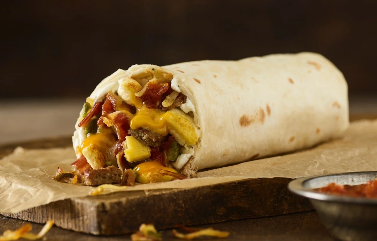 A wrapped burrito with a portion of the filling showing, containing meat, cheese, vegetables, and sauce, placed on a wooden surface with a bowl of salsa nearby. Food Styled in Denver, Colorado by Beth Hawkins of Hawkins and Company