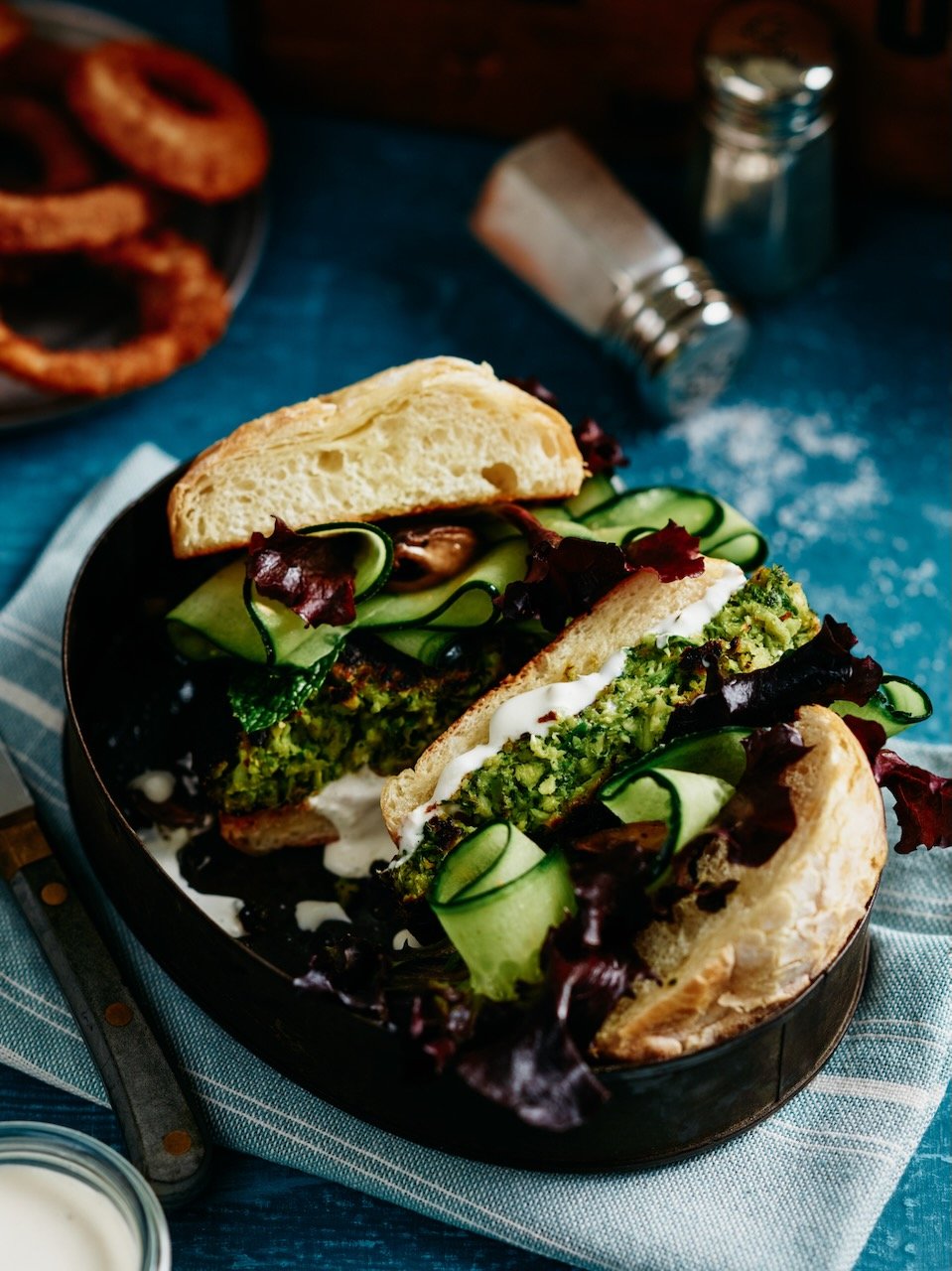 Vegan sandwich with avocado, cucumbers, lettuce, and creamy sauce on a toasted bun, served on a black plate.