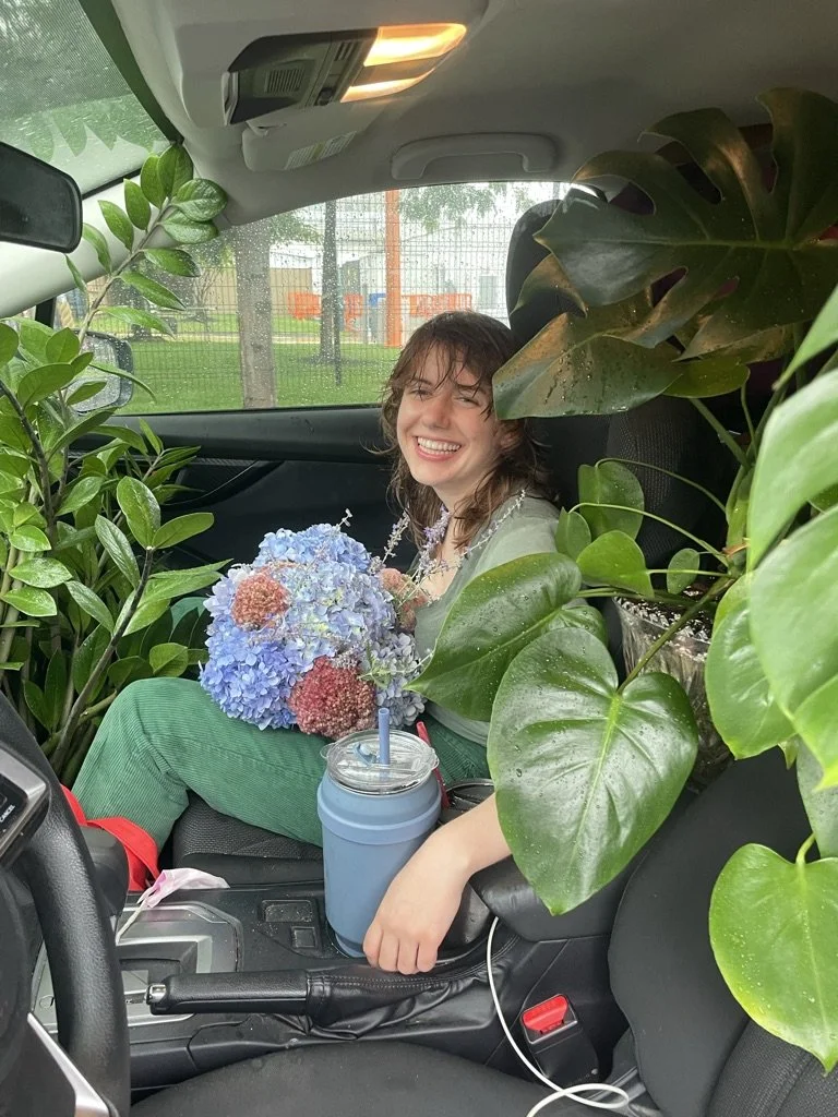 Alex, sitting in the passenger seat of a car, smiles while surrounded by lush plants and holding a bouquet of hydrangeas. She is on her way to a plant styling installation for a client, with plants filling the car