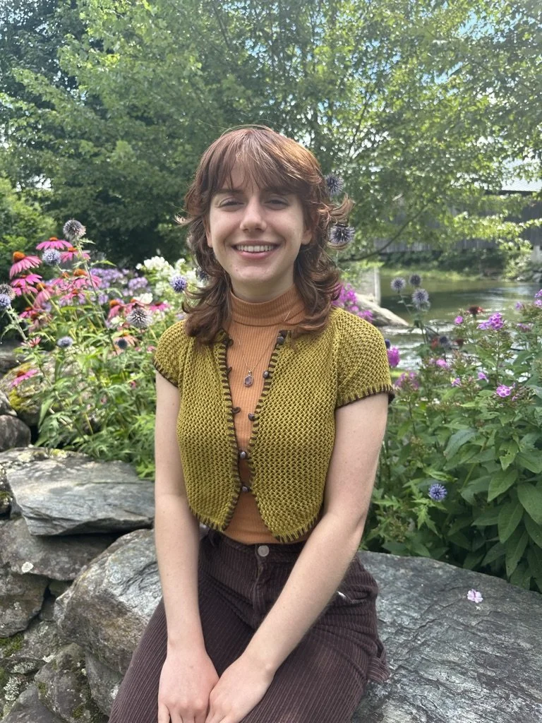 A woman with shoulder-length reddish-brown hair sits outdoors, smiling warmly. She is wearing a mustard yellow knit cardigan over a rust-colored turtleneck and brown pants. Behind her is a lush garden with colorful flowers and greenery, creating a pe