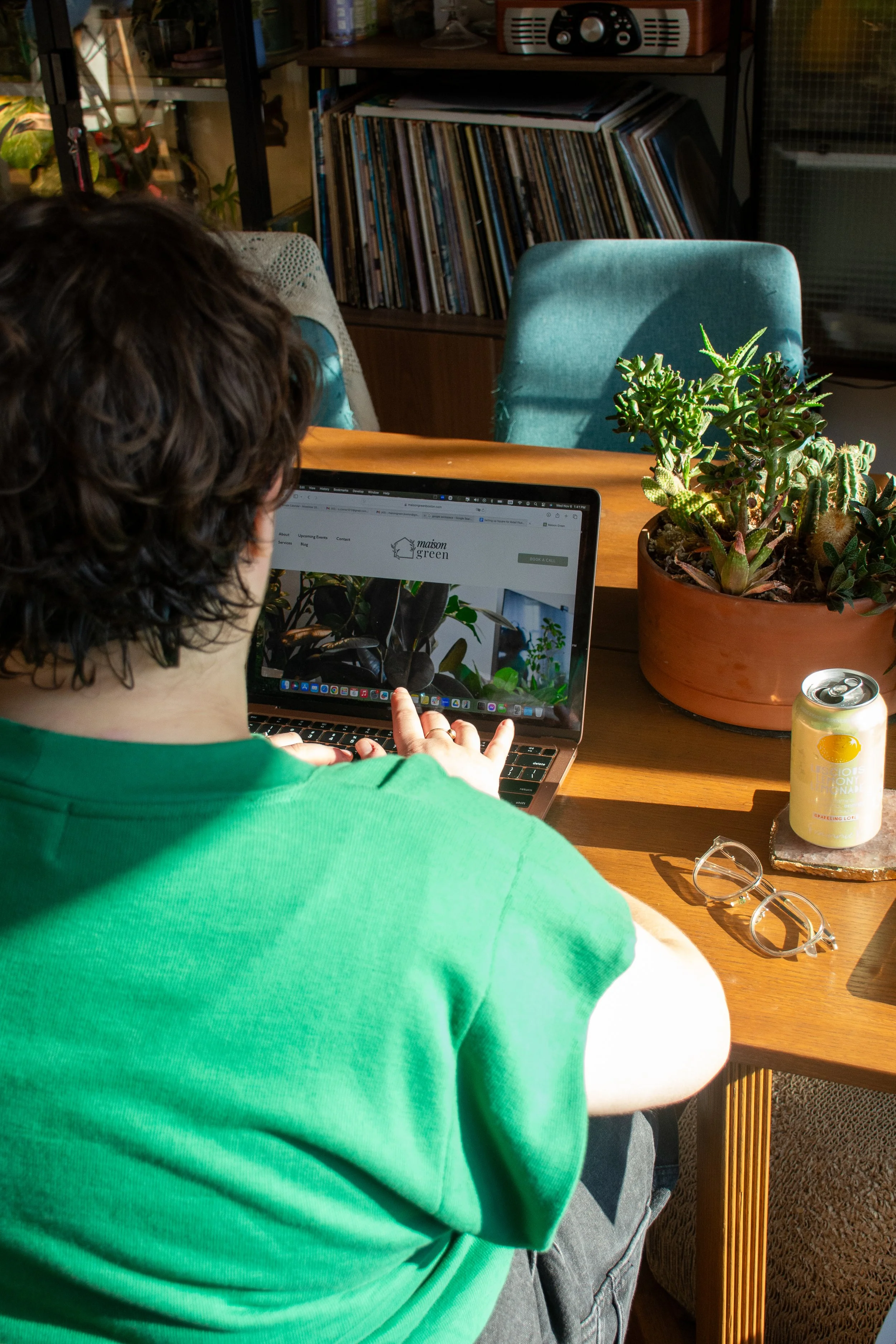 Sarah writing a personalized plant care report on her laptop at a table, surrounded by a potted succulent, a drink can, and glasses, bathed in natural light.