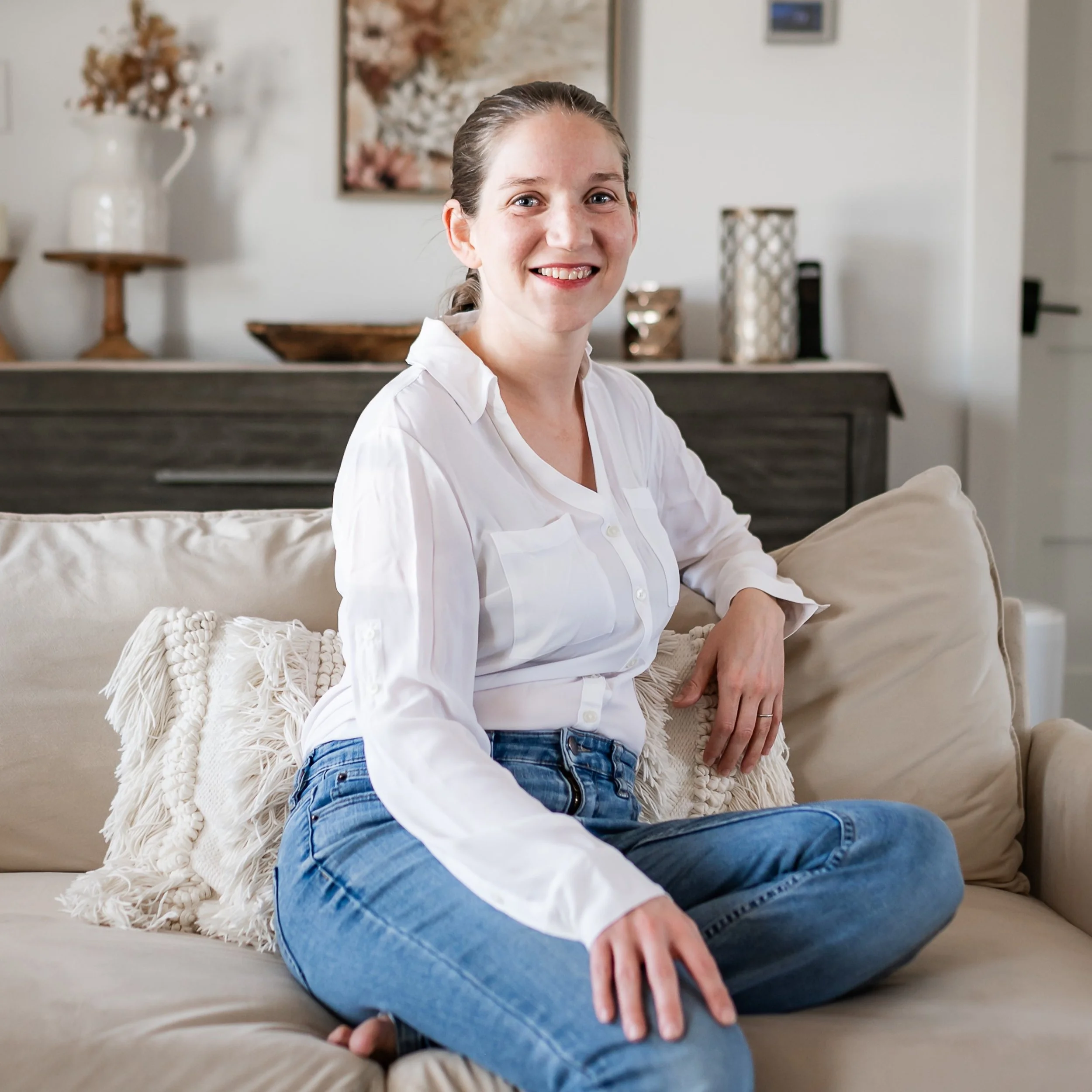 A woman with braided hair, wearing a white shirt and blue jeans, sitting on a beige sofa in a well-lit living room, smiling at the camera.