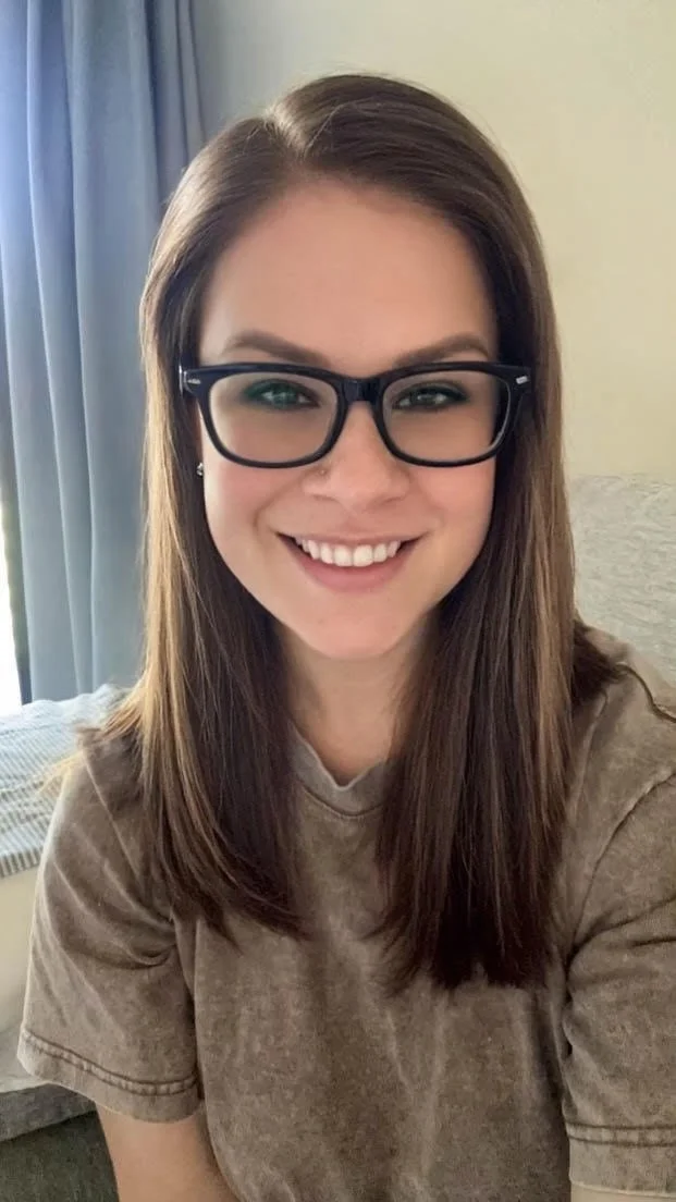 A woman with shoulder-length brown hair wearing glasses, smiling at the camera, in a room with light-colored walls, a window with curtains, and a bed in the background.