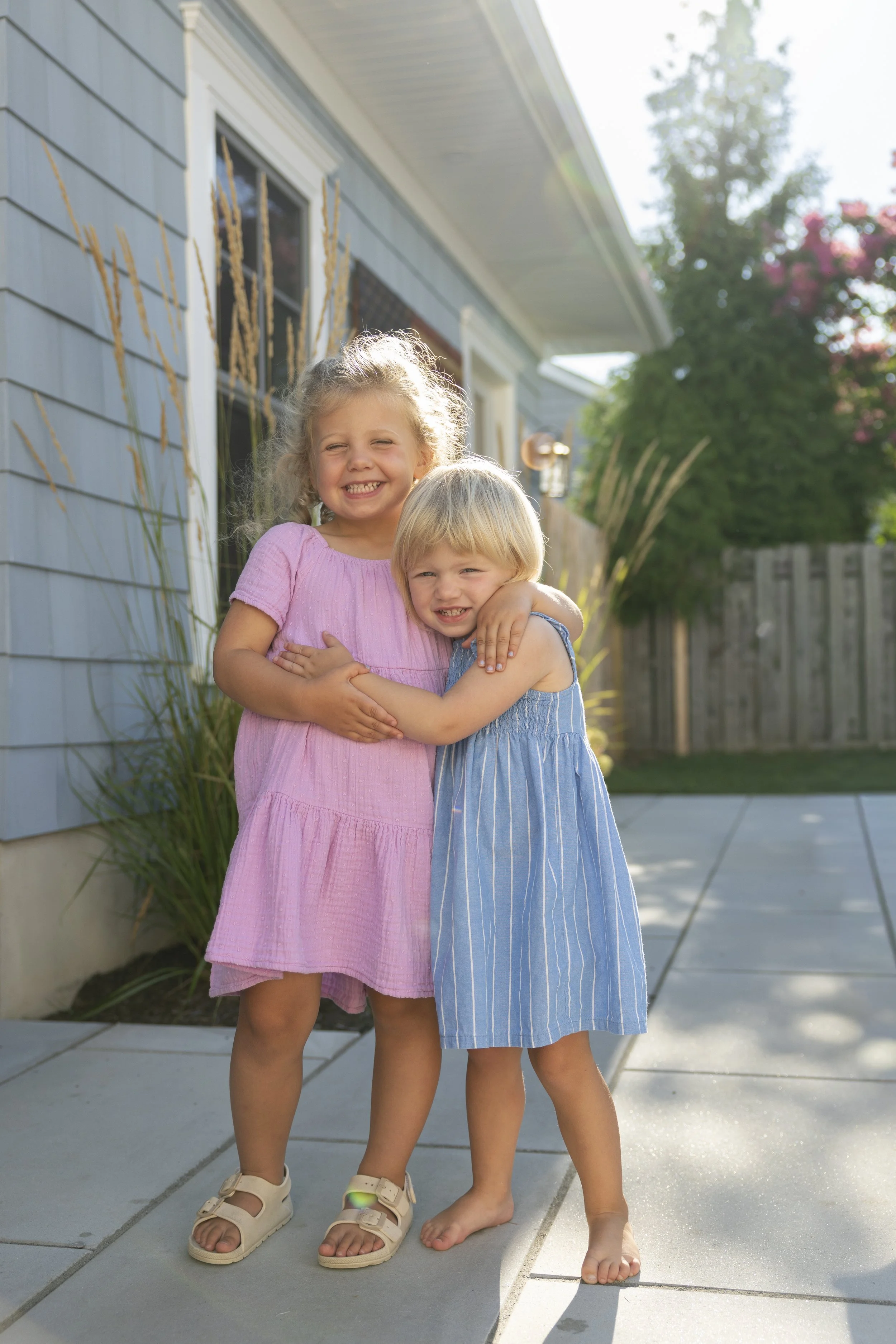 Two young sisters hugging each other and smiling outside near a light blue house, with a tree and a fenced yard in the background, sunlight shining down.