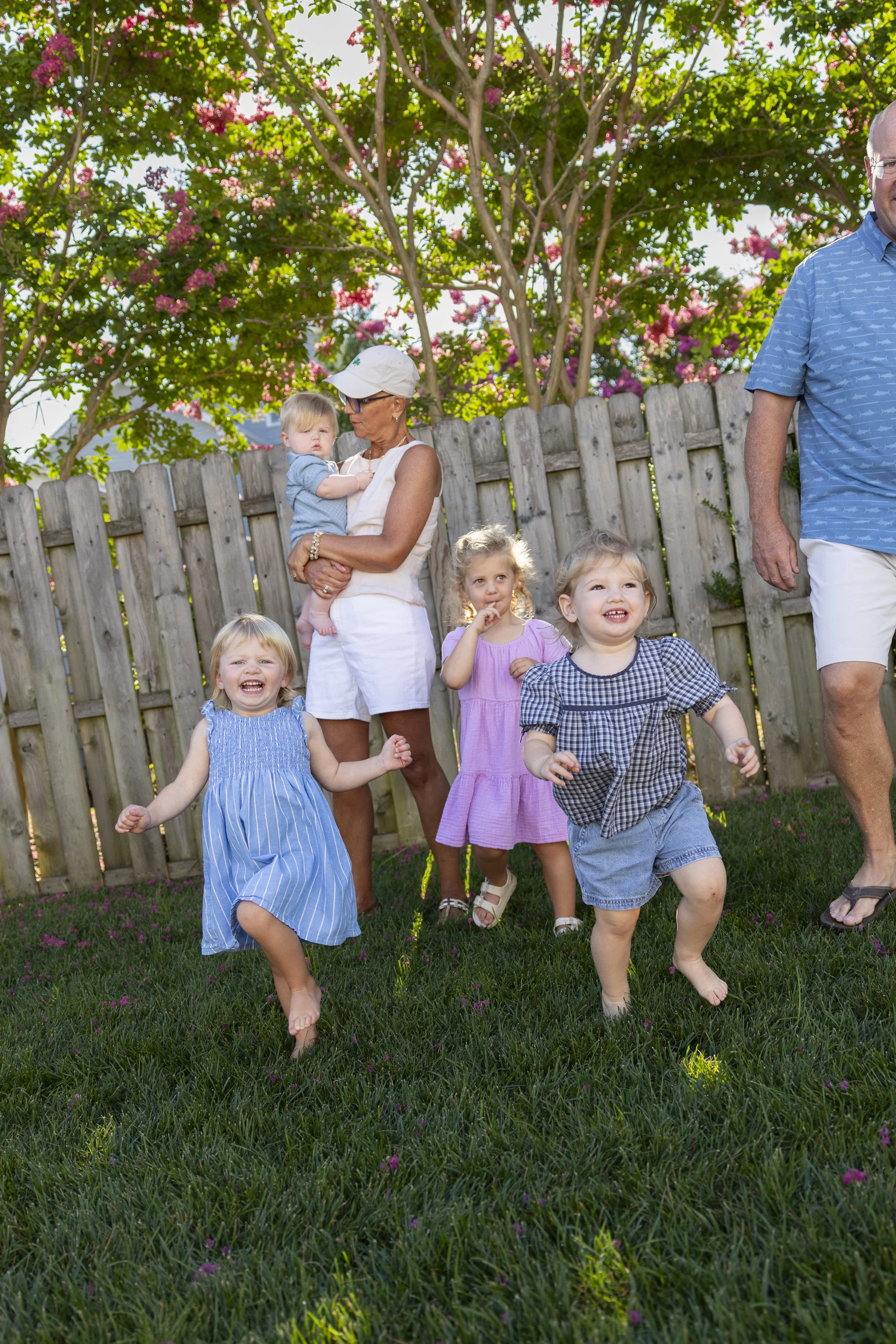 A group of children and adults playing outdoors on a grassy yard with a wooden fence and trees with pink flowers in the background. The children are happily running and smiling.