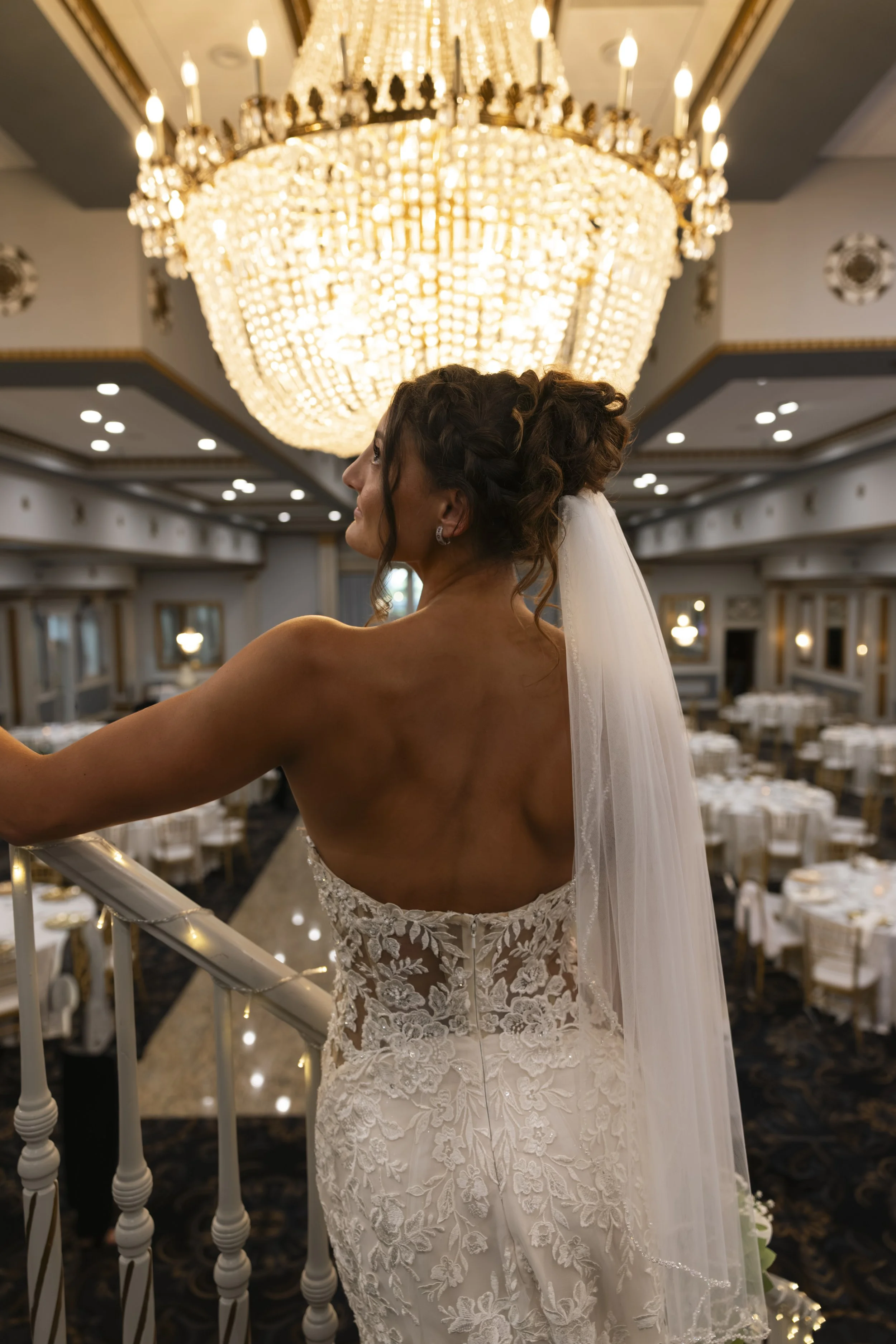 A bride in a strapless white lace wedding dress with a veil standing on a balcony in a grand ballroom with a large chandelier and decorated tables.