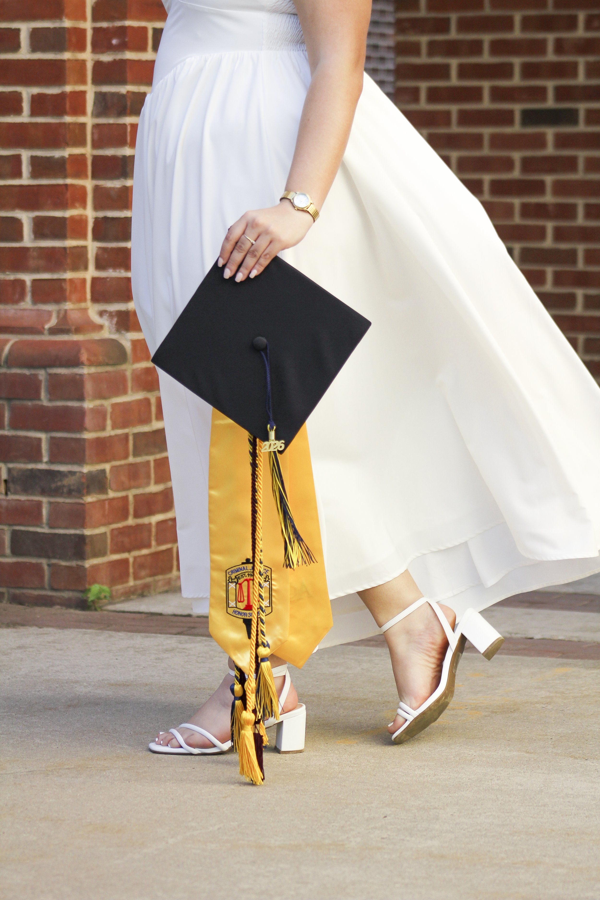 A person dressed in a graduation gown and cap is walking outside, wearing white high-heeled sandals and holding a black scholarship cap with a tassel.