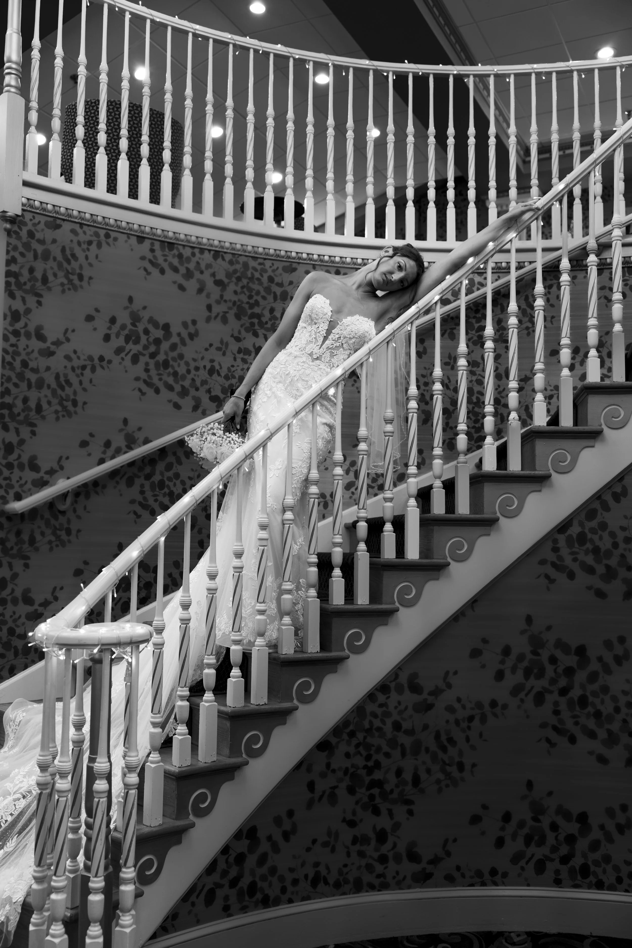 A woman in a wedding dress is leaning on a staircase railing, holding a bouquet of flowers, in an indoor setting with decorative background.