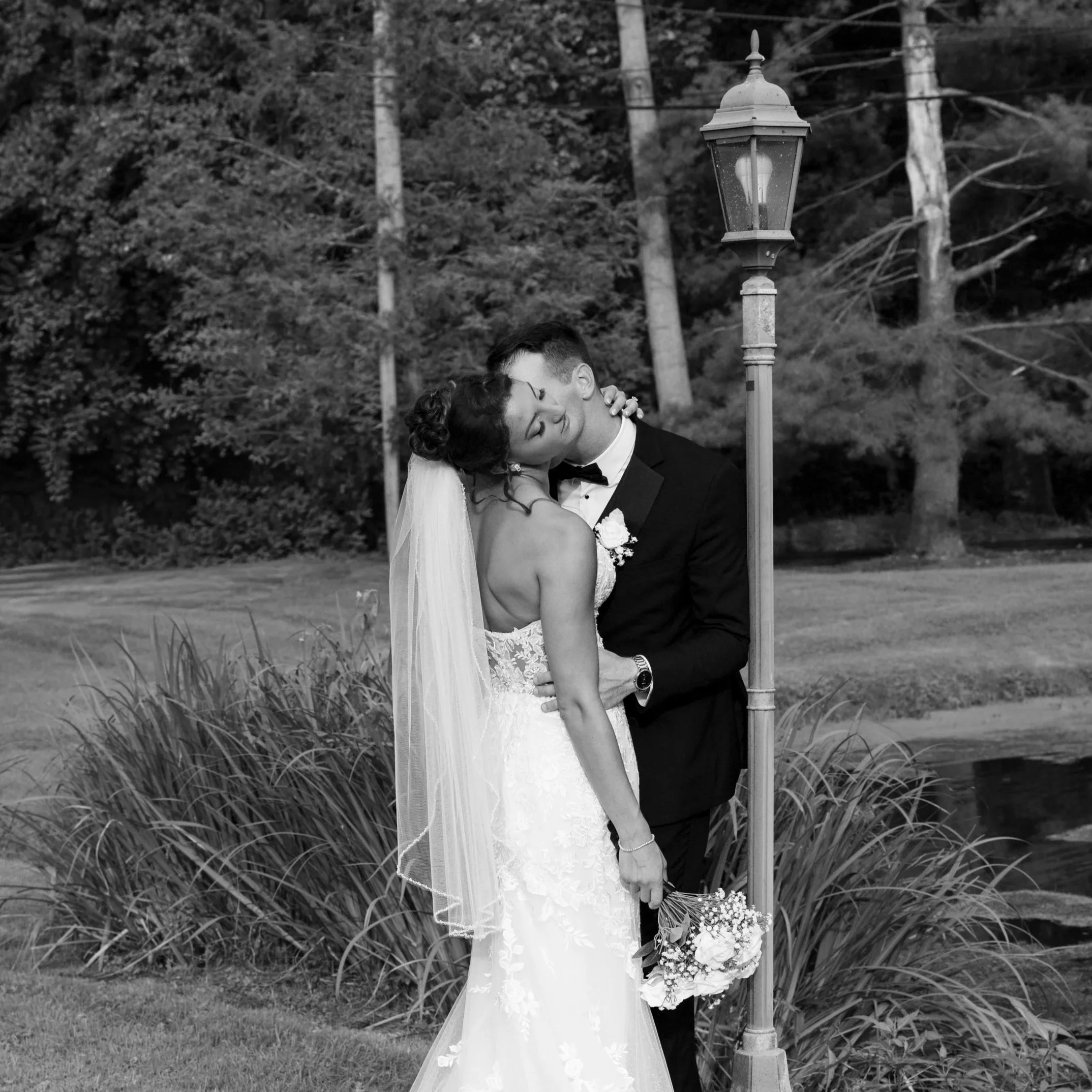 A bride and groom kissing outdoors during their wedding, with a lamppost and trees in the background, in black and white.