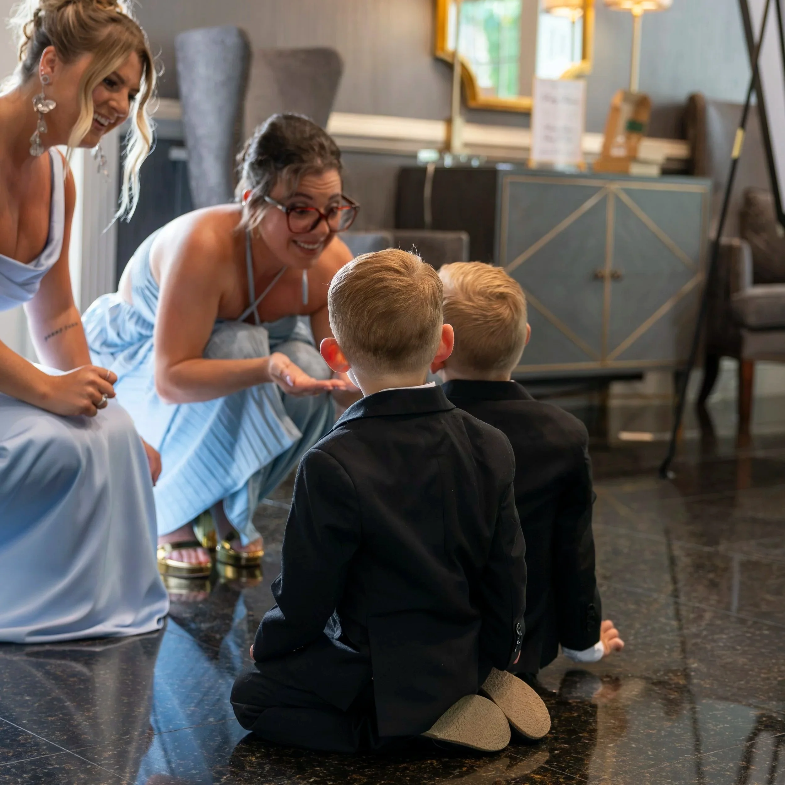 Two women and two young children crouch on a shiny, black floor, engaging in a playful activity in a well-lit room with decorative storage furniture and large windows.
