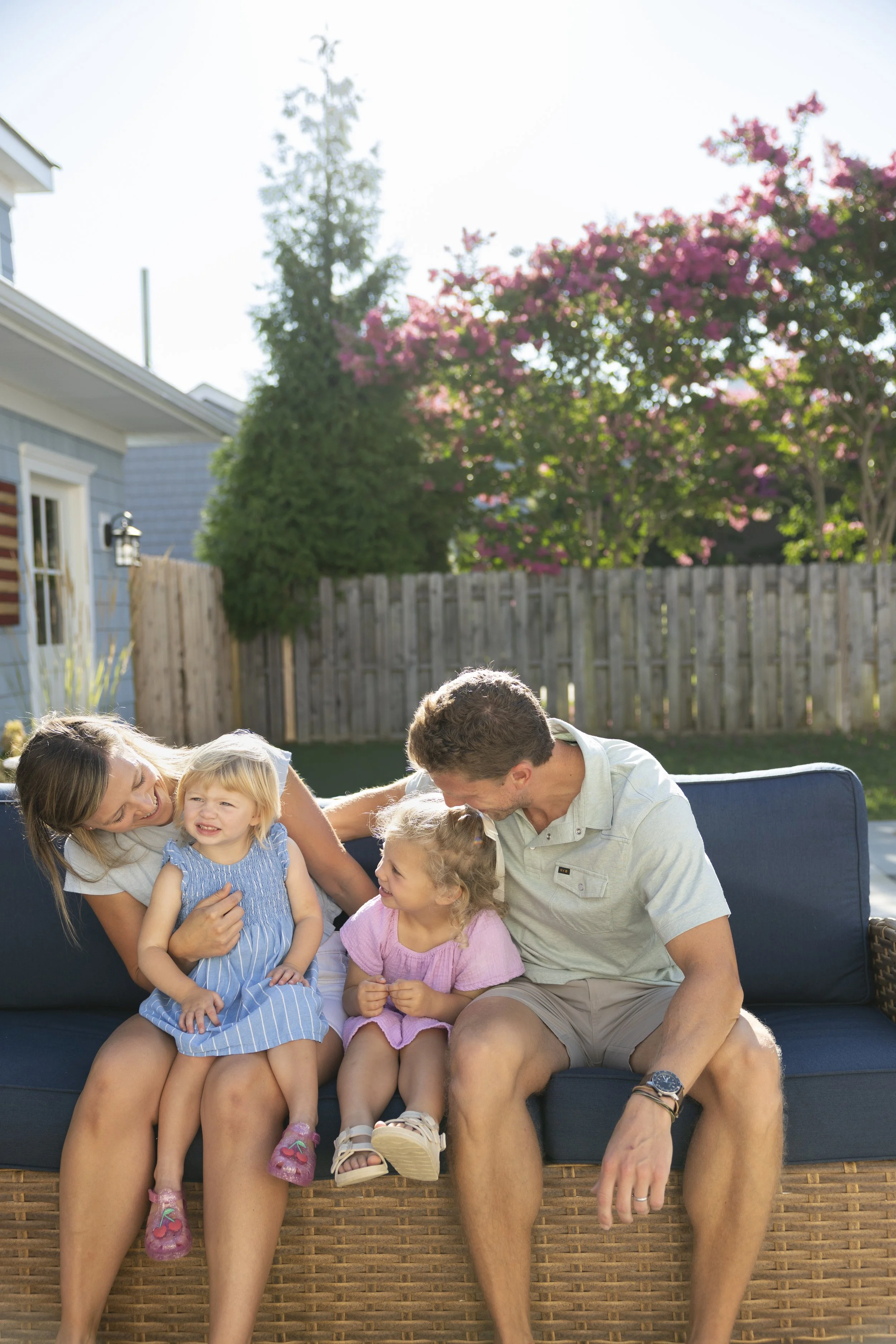 A family sitting outdoors on a couch in a backyard with pink blooming trees and a wooden fence in the background. The family includes two women, two young girls, and a man, all smiling and engaging happily.