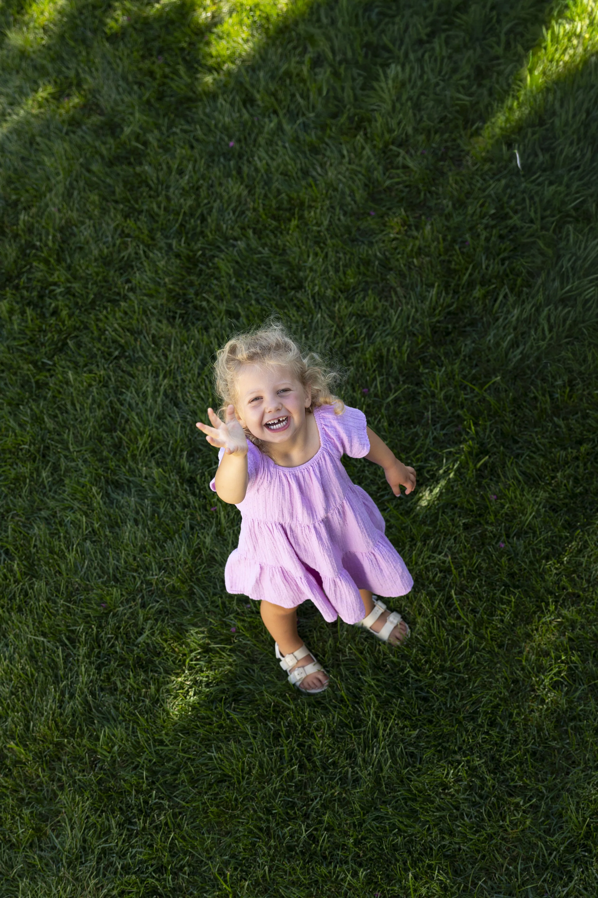 A young girl with curly blonde hair wearing a pink dress and white sandals is looking up and smiling while standing on green grass in a park, with her hand raised towards the camera.