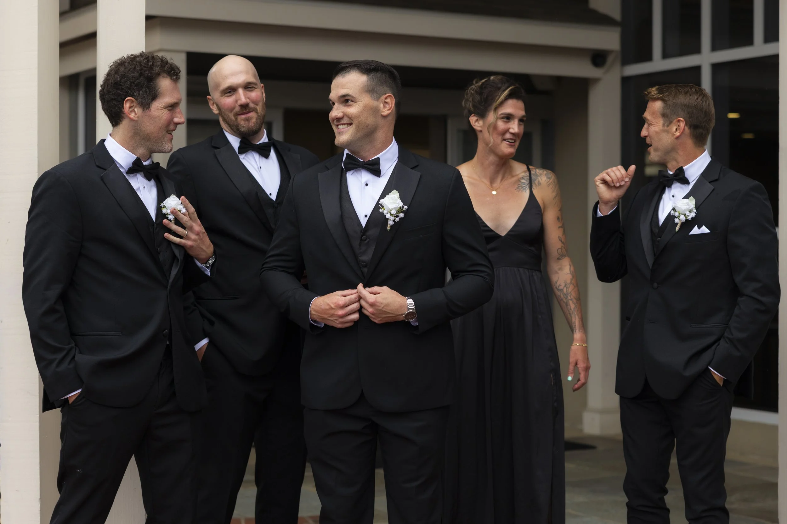 The groom and his siblings dressed in black tuxedos and one woman in a black dress talking and smiling outside a building