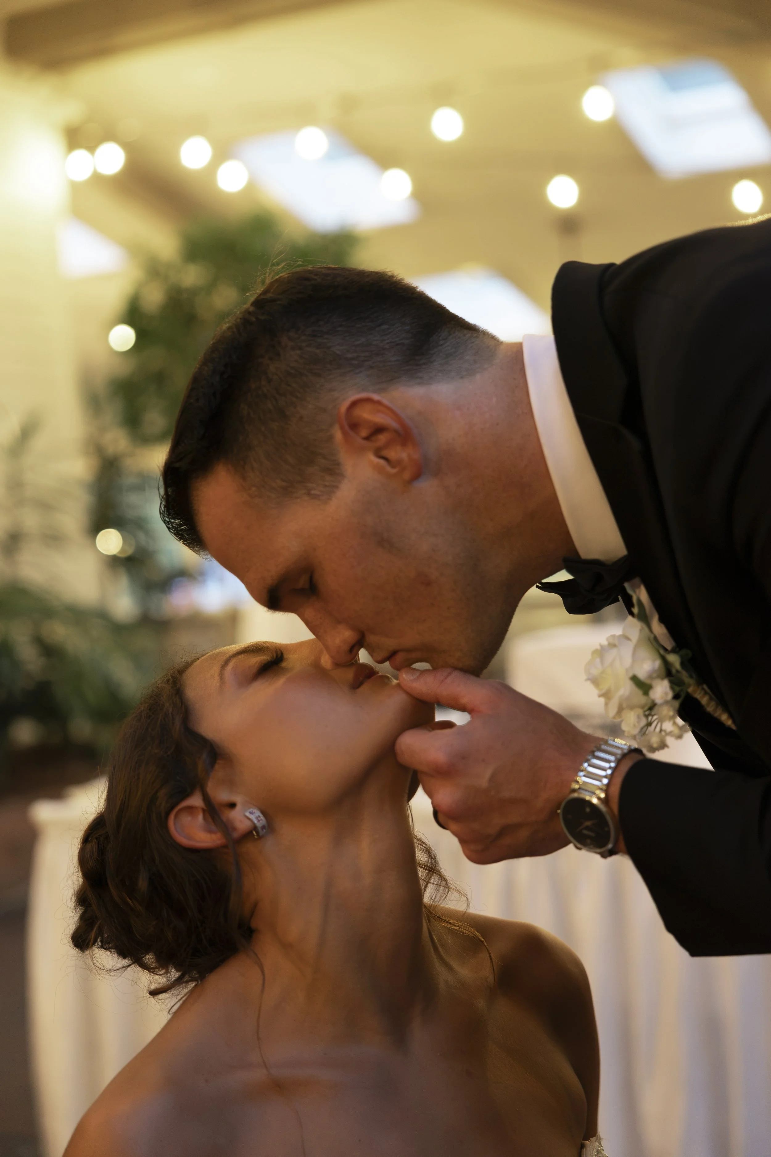 A man in a tuxedo shares an intimate moment with a woman, possibly during a wedding or special event, in a warmly lit indoor setting.